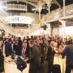 An elegant indoor event with people mingling and talking, featuring opulent balconies and ornate metalwork.