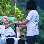 Young girl holding hands with elderly man in a wheelchair outdoors, surrounded by greenery.