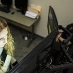 Two women in an office setting; one focused on a computer screen, the other wearing vr goggles and headphones.