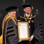 Two people in academic regalia hold a framed citation in front of a University of Central Florida backdrop.